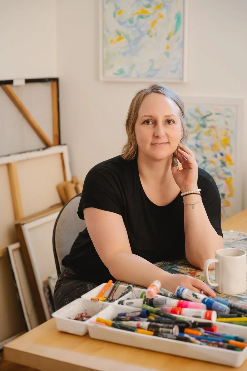 A woman sitting at a table with various art supplies, including markers and paints, in an art studio with paintings on the wall behind her.