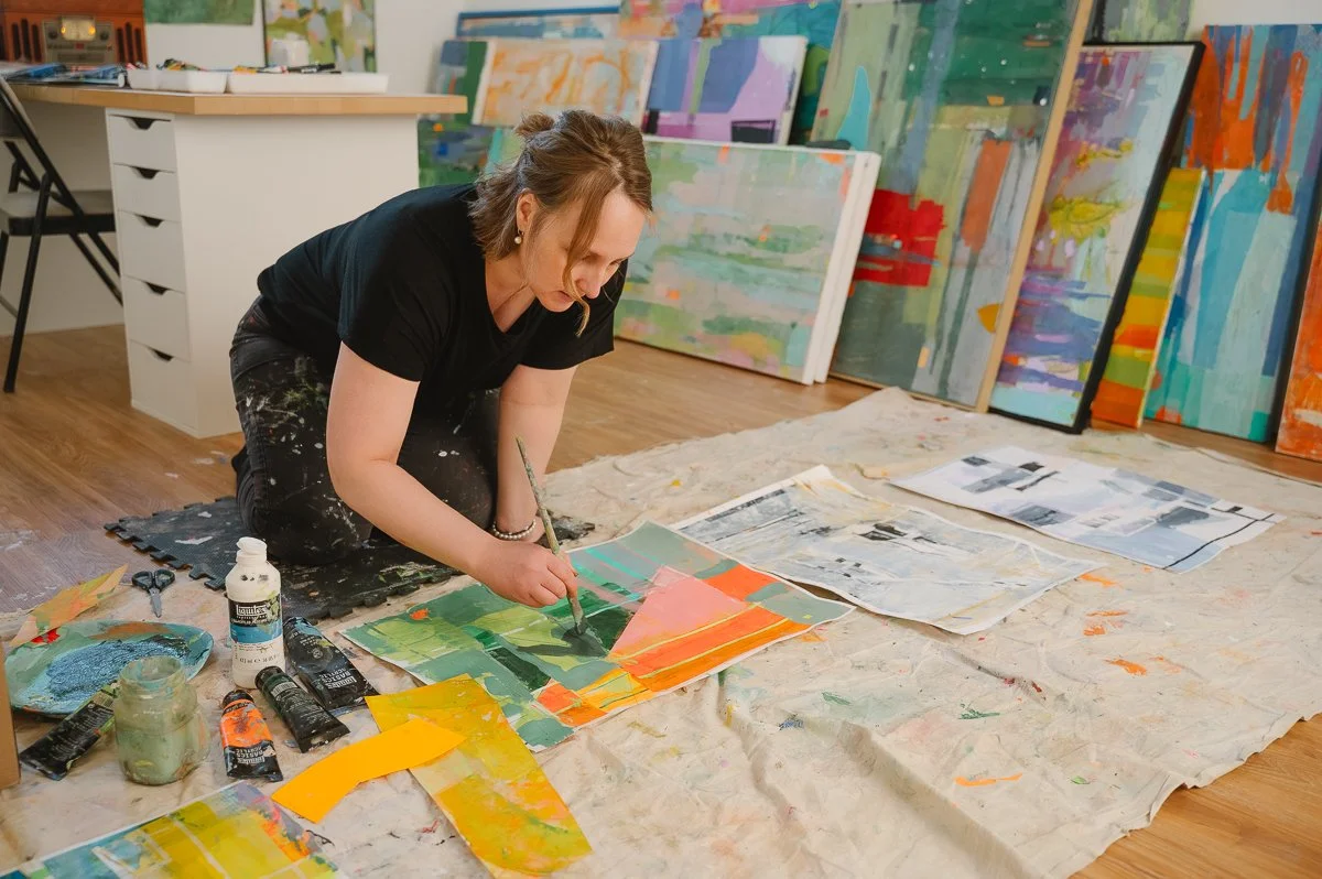 A woman kneeling on the floor, painting an abstract landscape on paper with vibrant colors, surrounded by completed paintings and art supplies in an art studio.