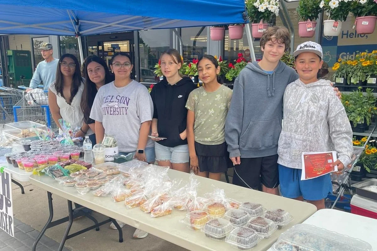 Group of children standing behind a table of baked goods at a bake sale outside a store.