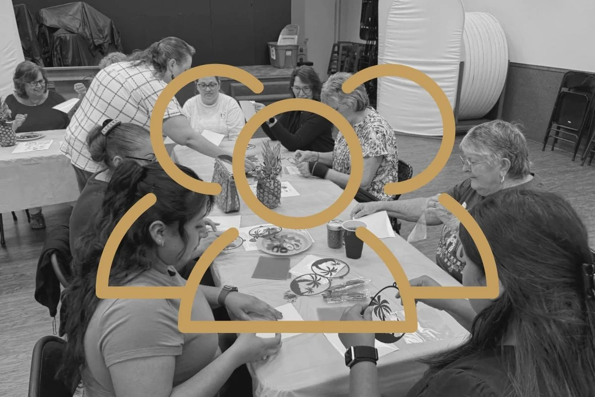 A group of women sitting around a table engaging in arts and crafts, with pineapple decorations on the table, in a room with stacked chairs and large objects in the background.