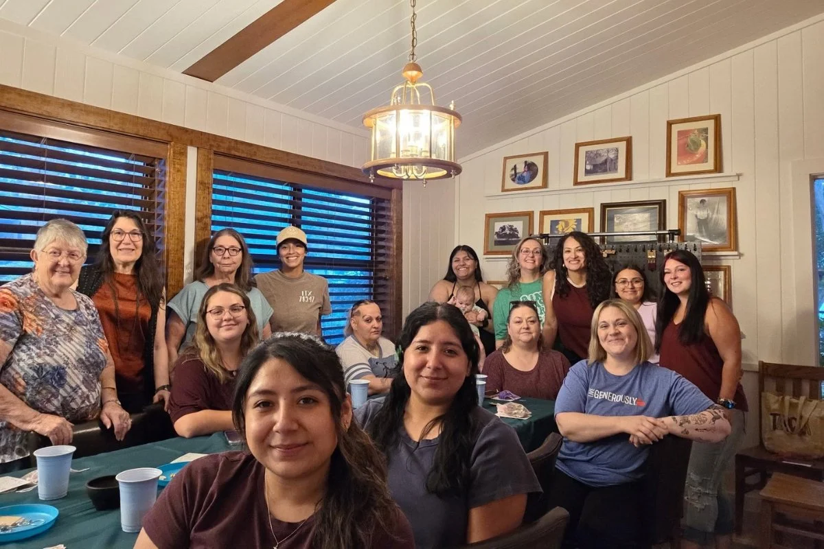 Group of women gathered around a dining table in a cozy room with framed pictures on the wall, inside a house.