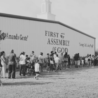 Line of people standing outside a building with a sign that reads 'First Assembly of God' and a quote from the Bible, John 3:16.