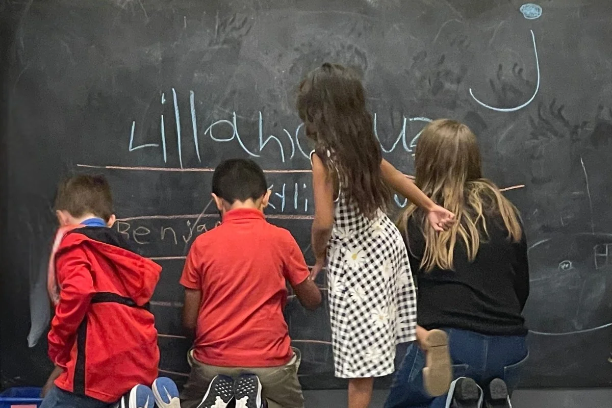 Four children sitting and standing in front of a blackboard, writing and drawing with chalk.