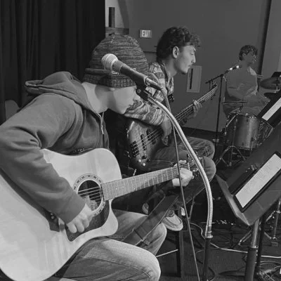 Three musicians playing guitars and drums in a practice space.