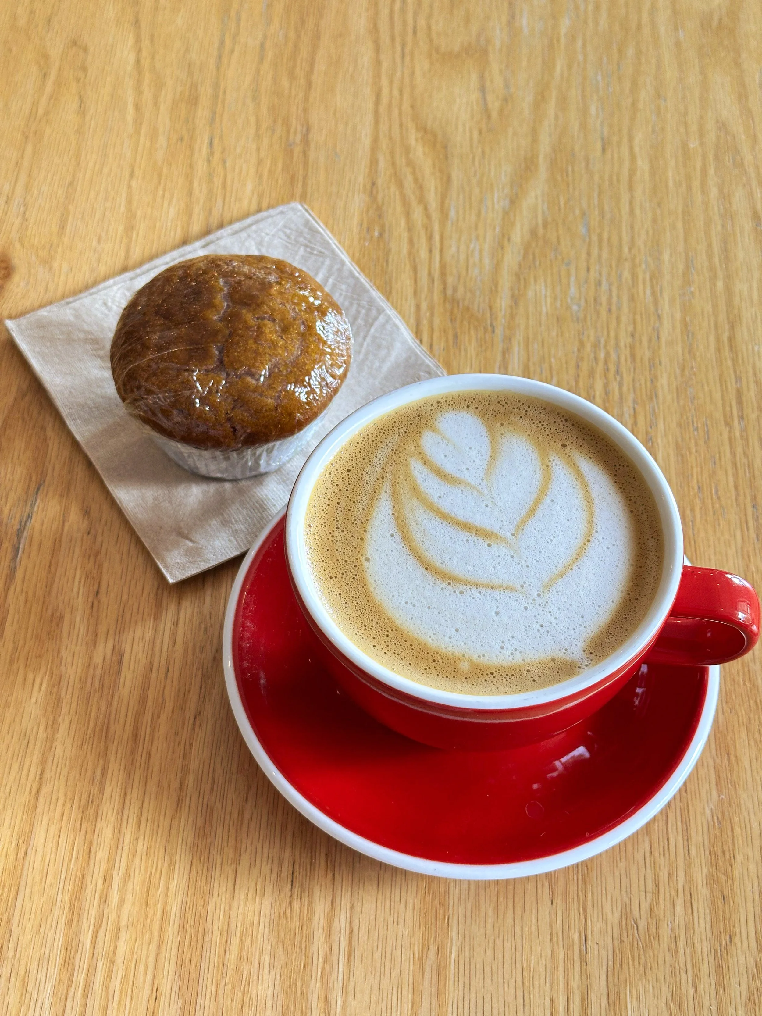 A cup of coffee with latte art in a red mug on a matching red saucer, and a muffin on a napkin on a wooden table.