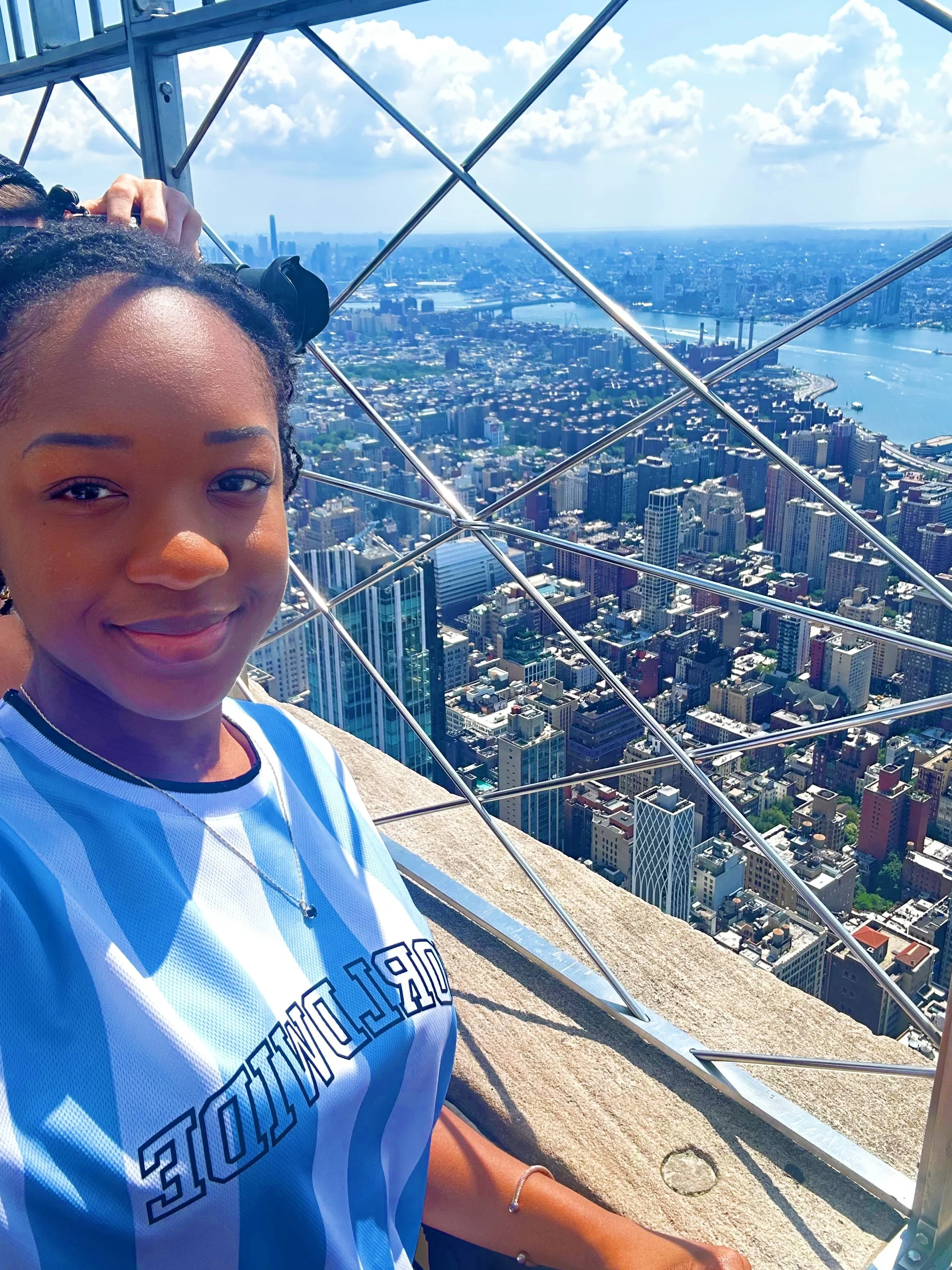 A young woman taking a selfie on a high observation deck with a cityscape view below. She is smiling, wearing a light blue and white sports jersey, and has her hair styled in braids. The city below features numerous tall buildings and a river.
