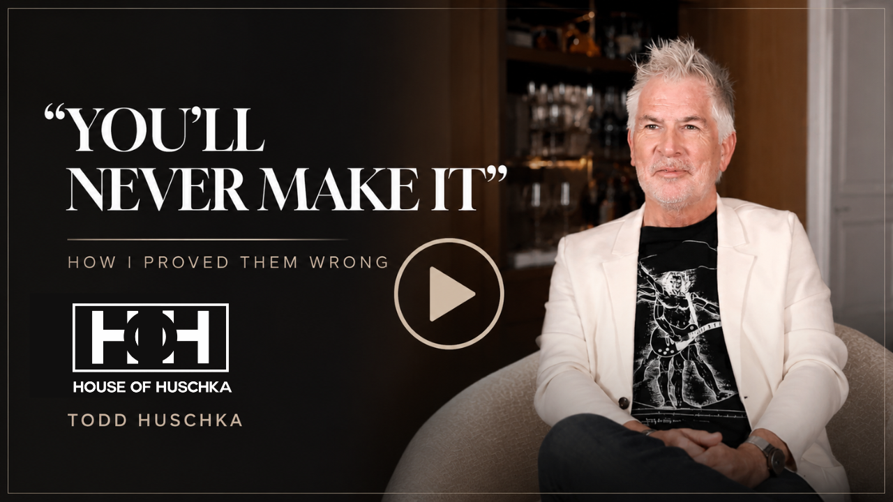 Promotion for a video titled 'You'll Never Make It' by Todd Huschka, featuring a man with spiky white hair wearing a white blazer and black graphic T-shirt, sitting on a beige couch in a dimly lit room with shelves of bottles in the background.
