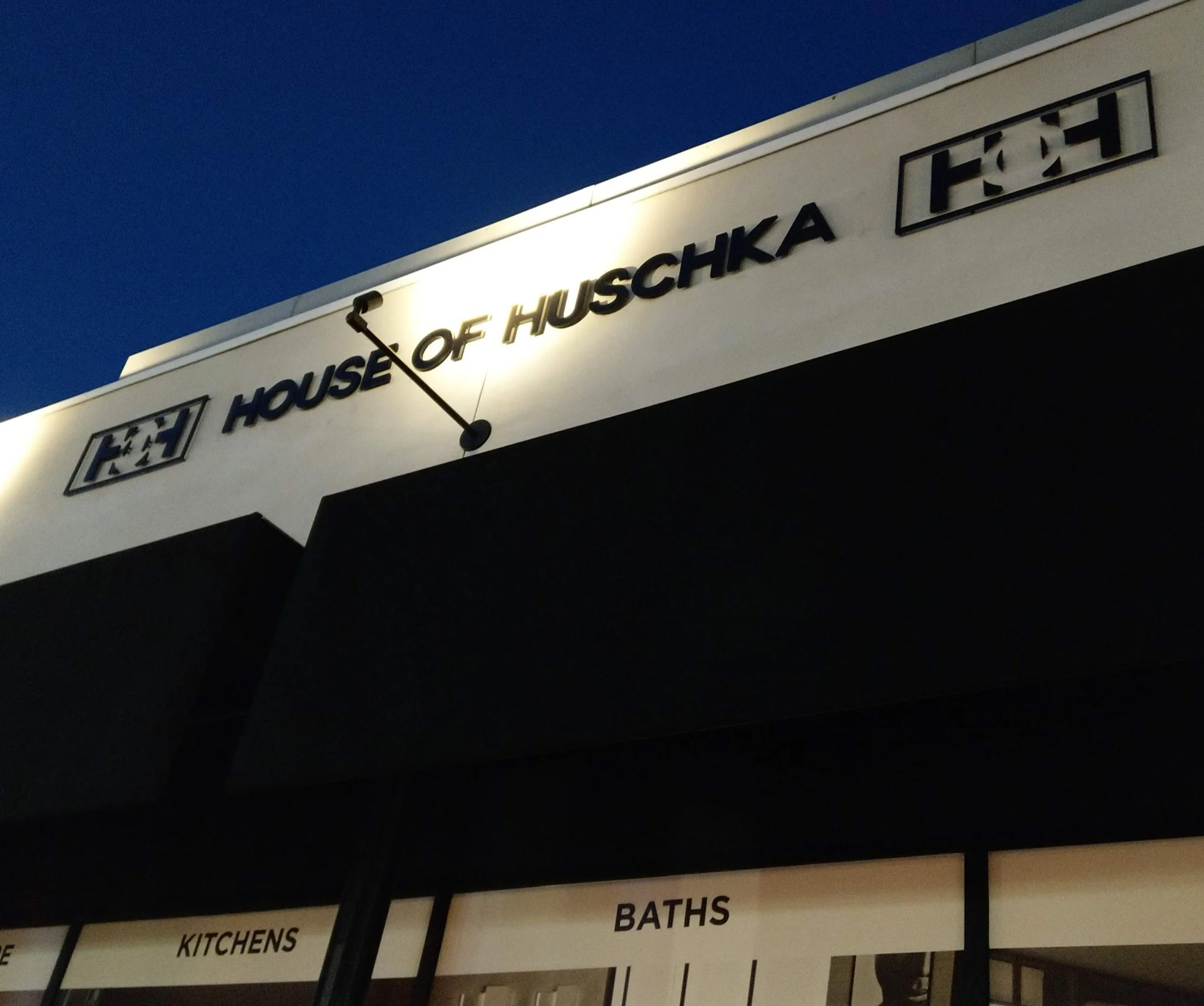 A sign on a building at dusk reads 'House of Hushka' and has directional signs for kitchens, baths, and other areas. The sky is dark blue.