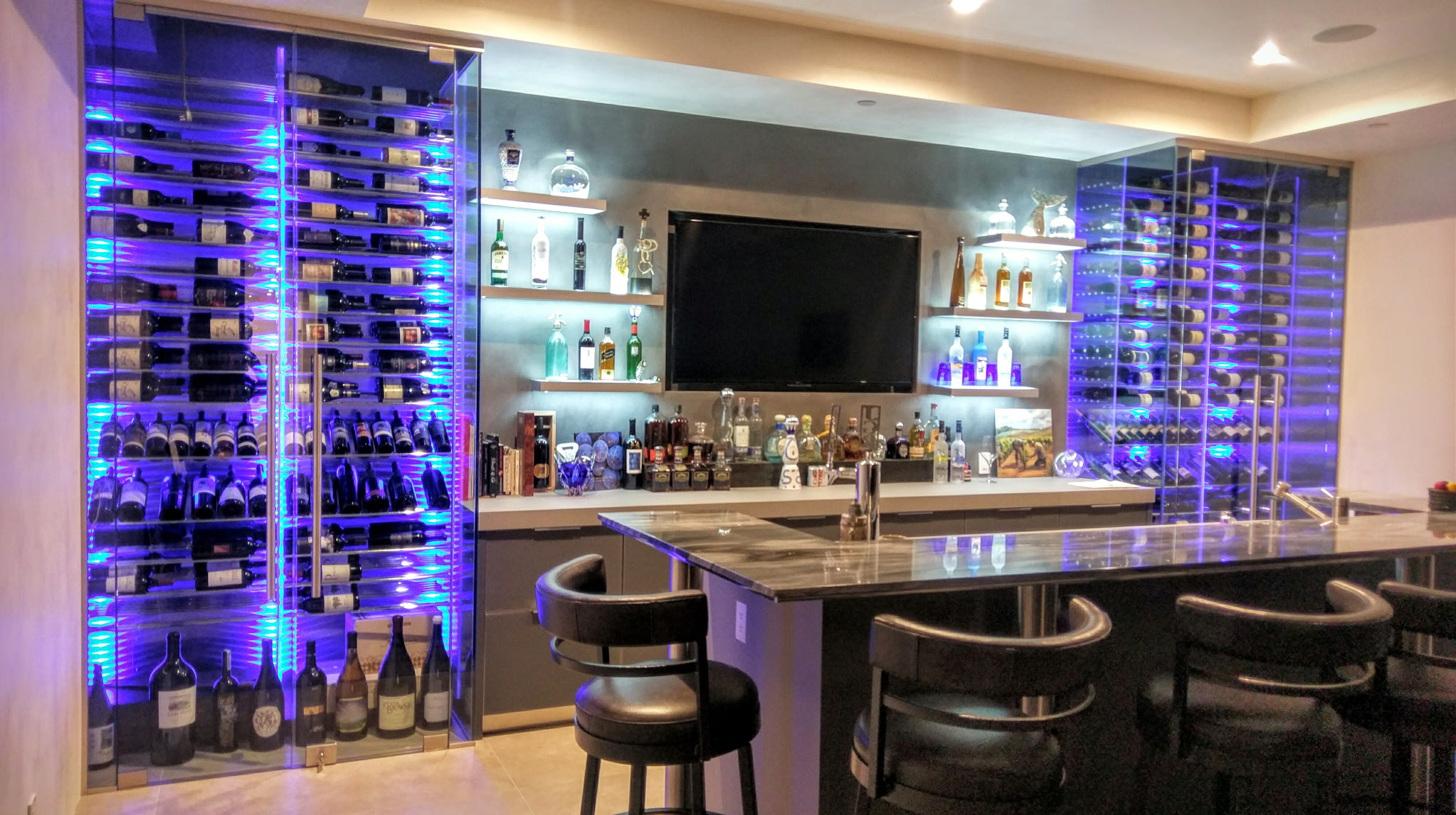 A home bar with illuminated wine storage, shelves of various liquor bottles, and a bar counter with stools, set in a modern style with ambient lighting.