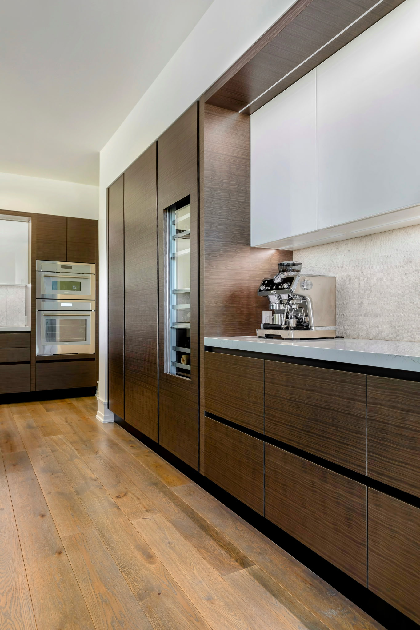 A modern kitchen with dark wood cabinets, a coffee machine on the countertop, and a built-in oven, with hardwood flooring.