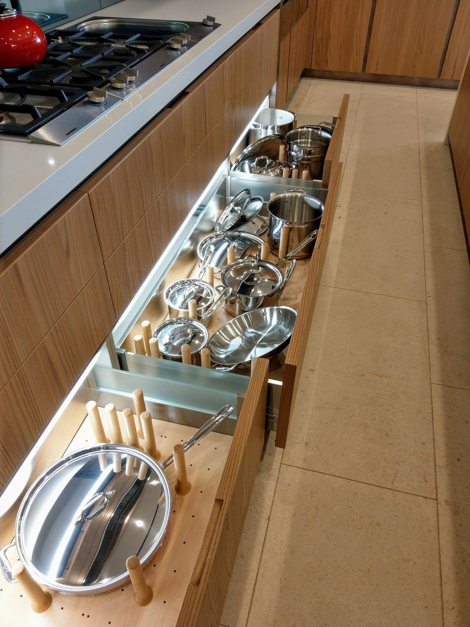 A kitchen countertop with a built-in gas stove and an open wooden drawer filled with stainless steel pots and pans, some with glass lids, and wooden-handled utensils.