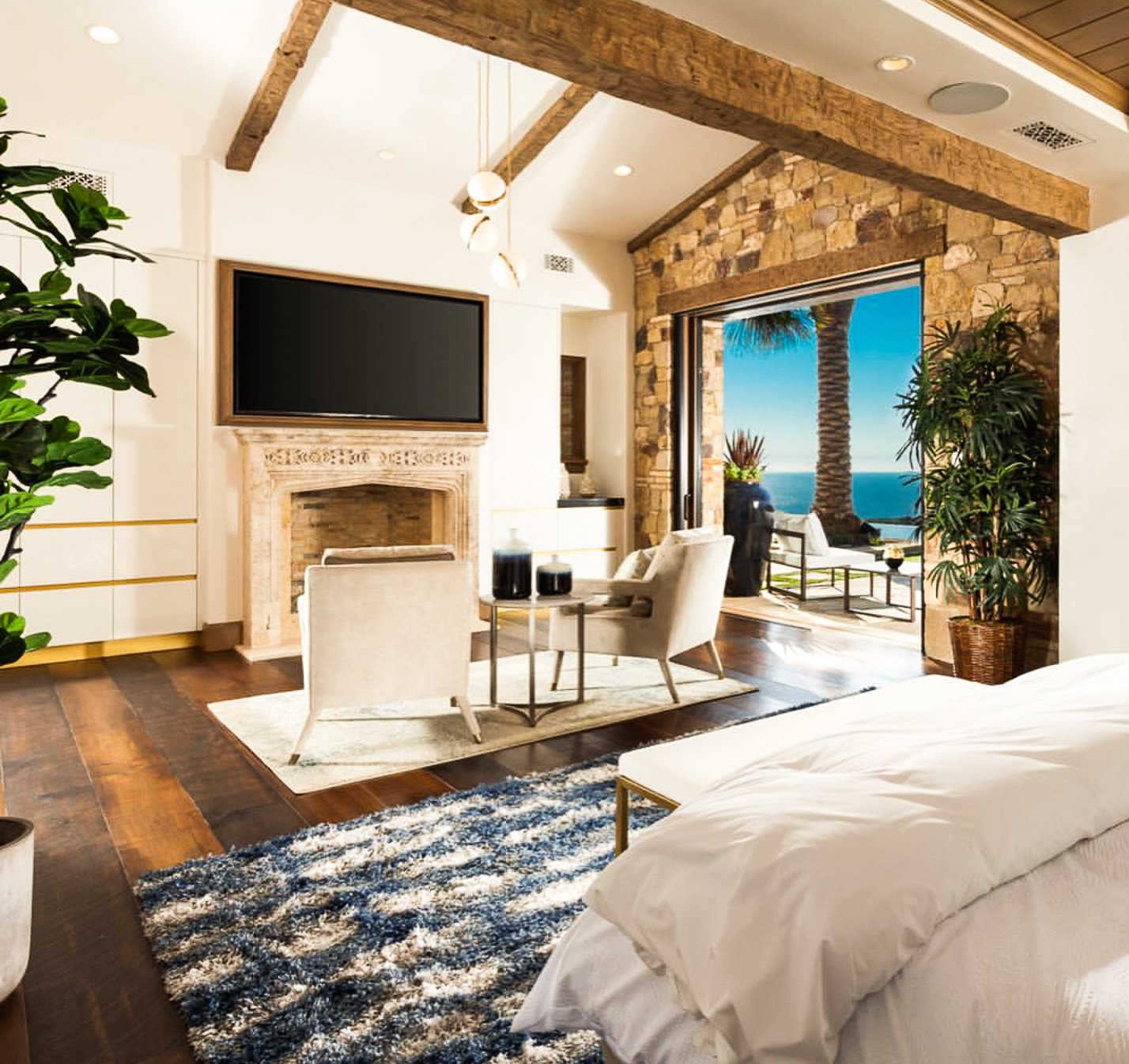 Living room with cream-colored furniture, a stone fireplace, a large flat-screen TV, plants, and an open sliding door leading to an outdoor seating area with a view of the ocean and palm trees.