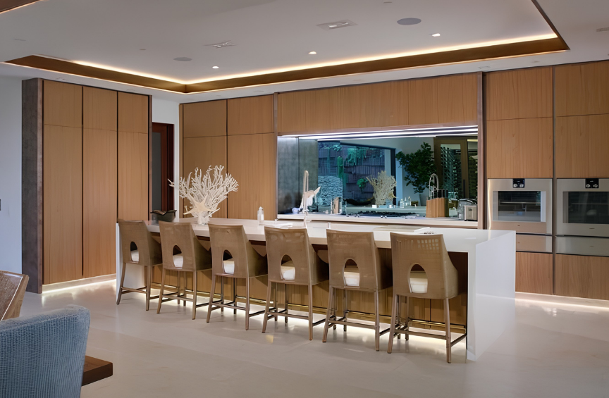Modern kitchen with wooden cabinets, white island counter, six beige chairs, large window with outdoor view, and built-in appliances.