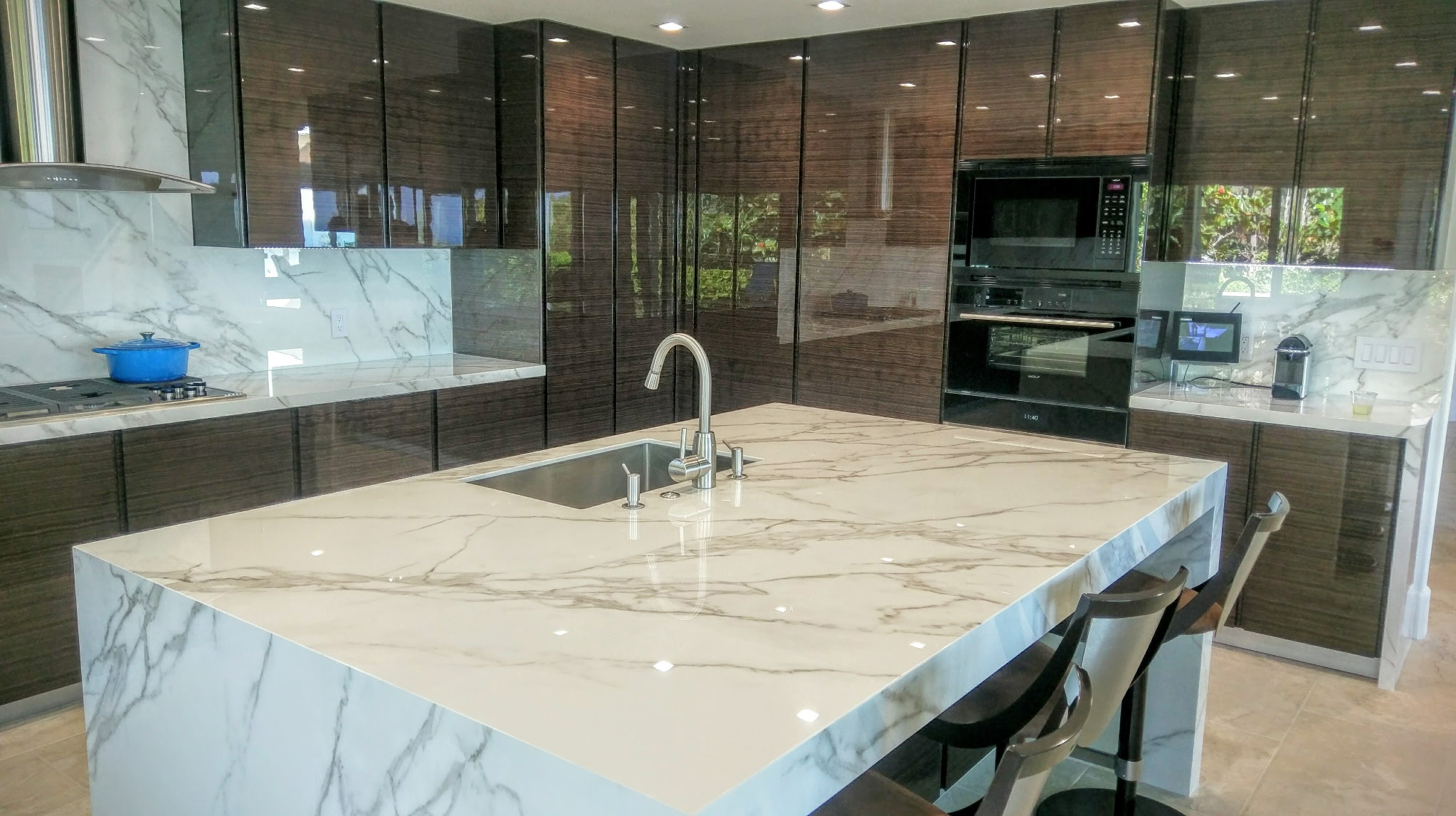 Modern kitchen with marble island, dark wood cabinets, built-in microwave, and view of greenery outside through reflections.