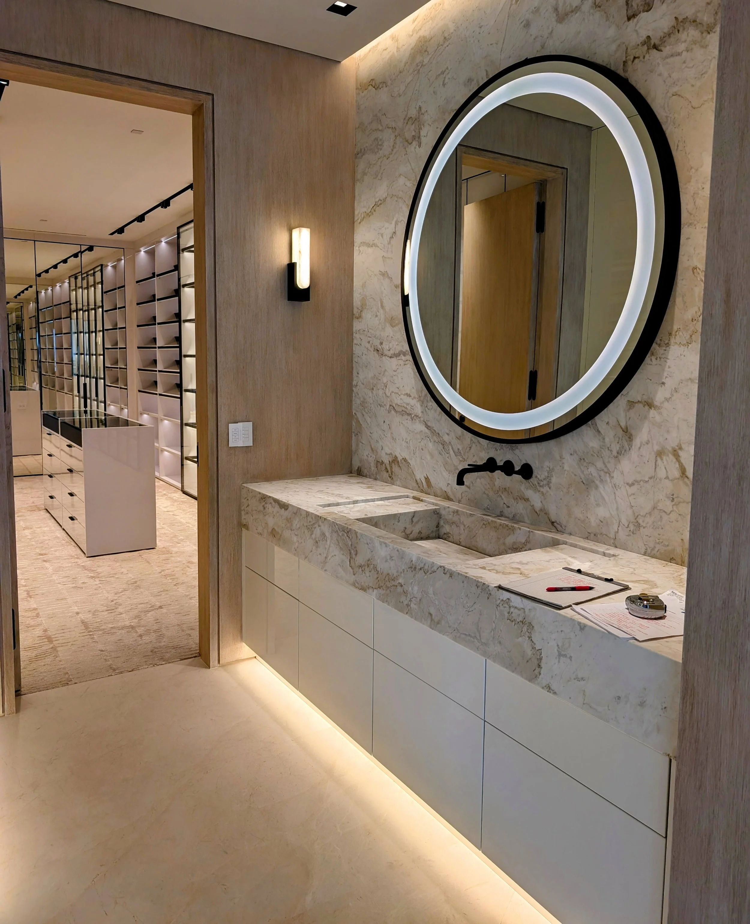 Modern bathroom with a circular backlit mirror above a marble countertop with a sink, a black faucet, and some papers and tools on the counter. There is a wall-mounted light fixture and beige floor and wall tiles. An open doorway reveals a hallway with wine racks or shelving.