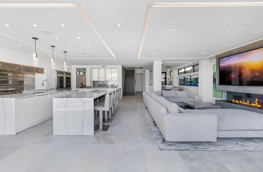 Modern open-concept kitchen and living room with white and gray color scheme, marble countertops, a fireplace, large television, and ample seating.