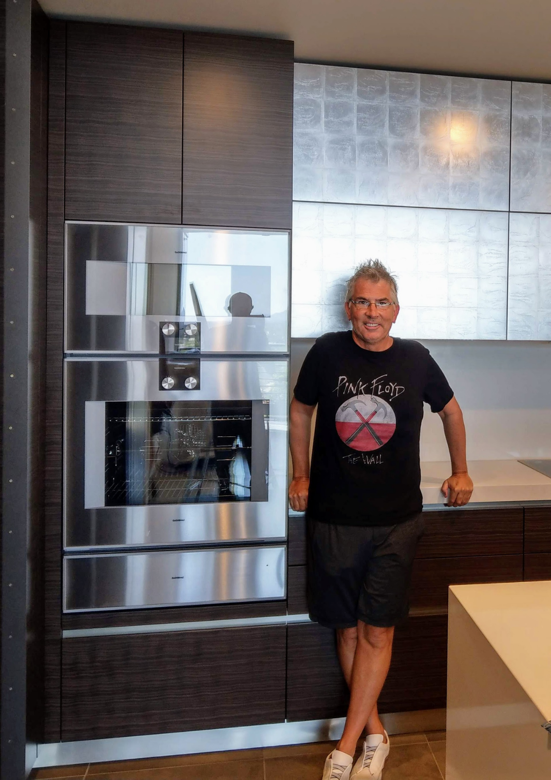 A man with gray hair and glasses standing in a modern kitchen. He is wearing a black Pink Floyd T-shirt, dark shorts, and white sneakers. He is smiling and has his arms resting on a kitchen counter.