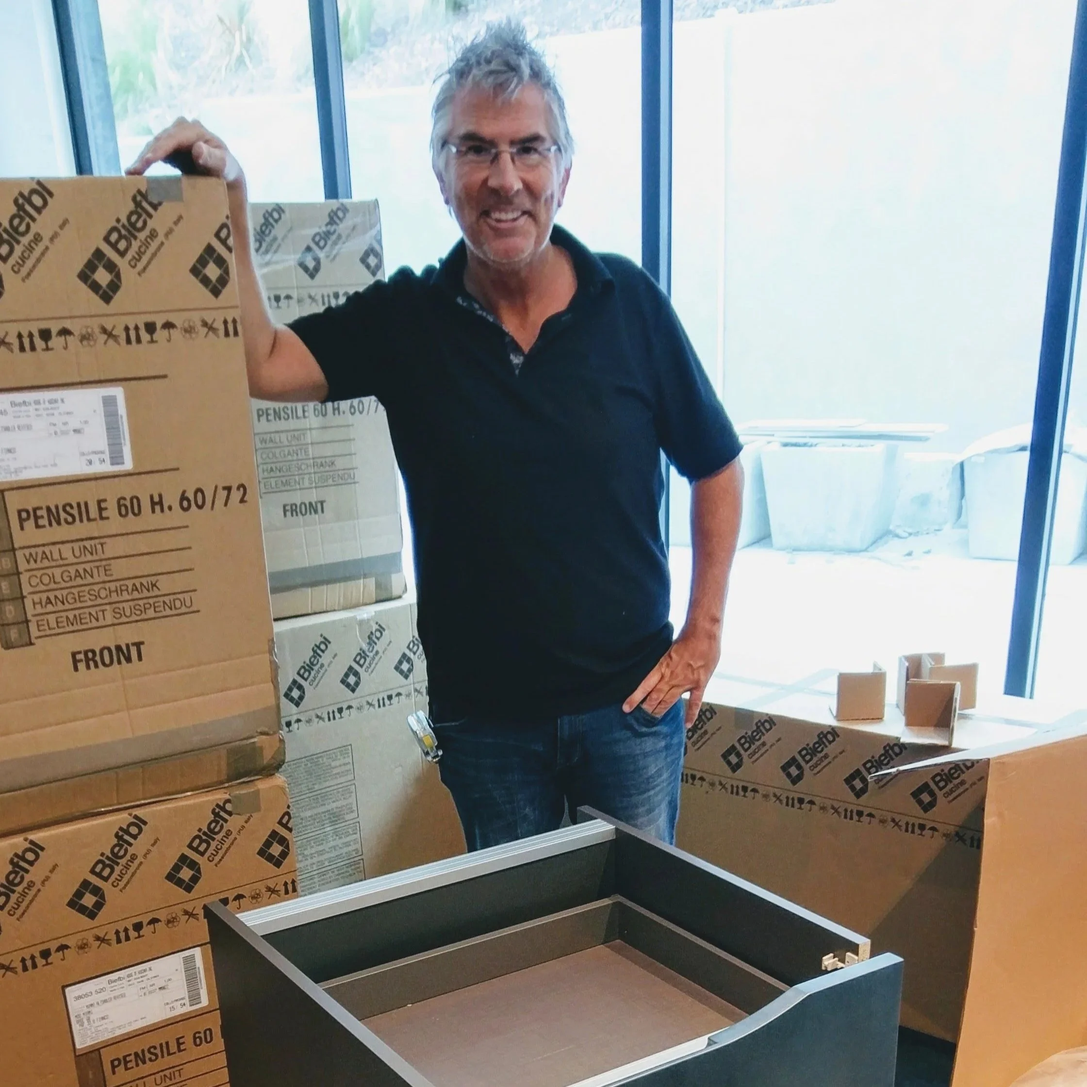 A man with gray hair, glasses, and a dark polo shirt smiling and holding a cardboard box inside a store or office with large windows, surrounded by other boxes.