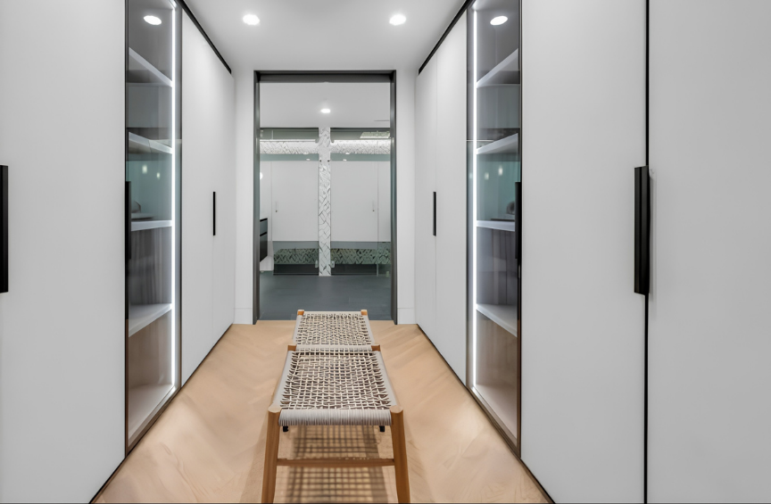 Modern walk-in closet with white cabinets, built-in shelving, and a bench in the center, leading to an adjacent room with glass doors.