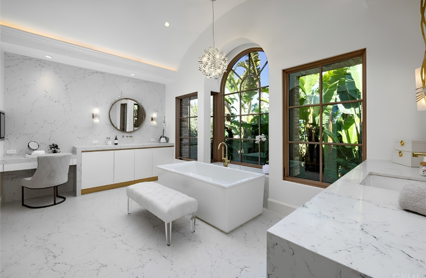 A luxurious bathroom with white marble floors and walls, a large built-in bathtub with a gold faucet, large window views of tropical plants, and elegant decor including a round mirror, chandelier, and vanity area.