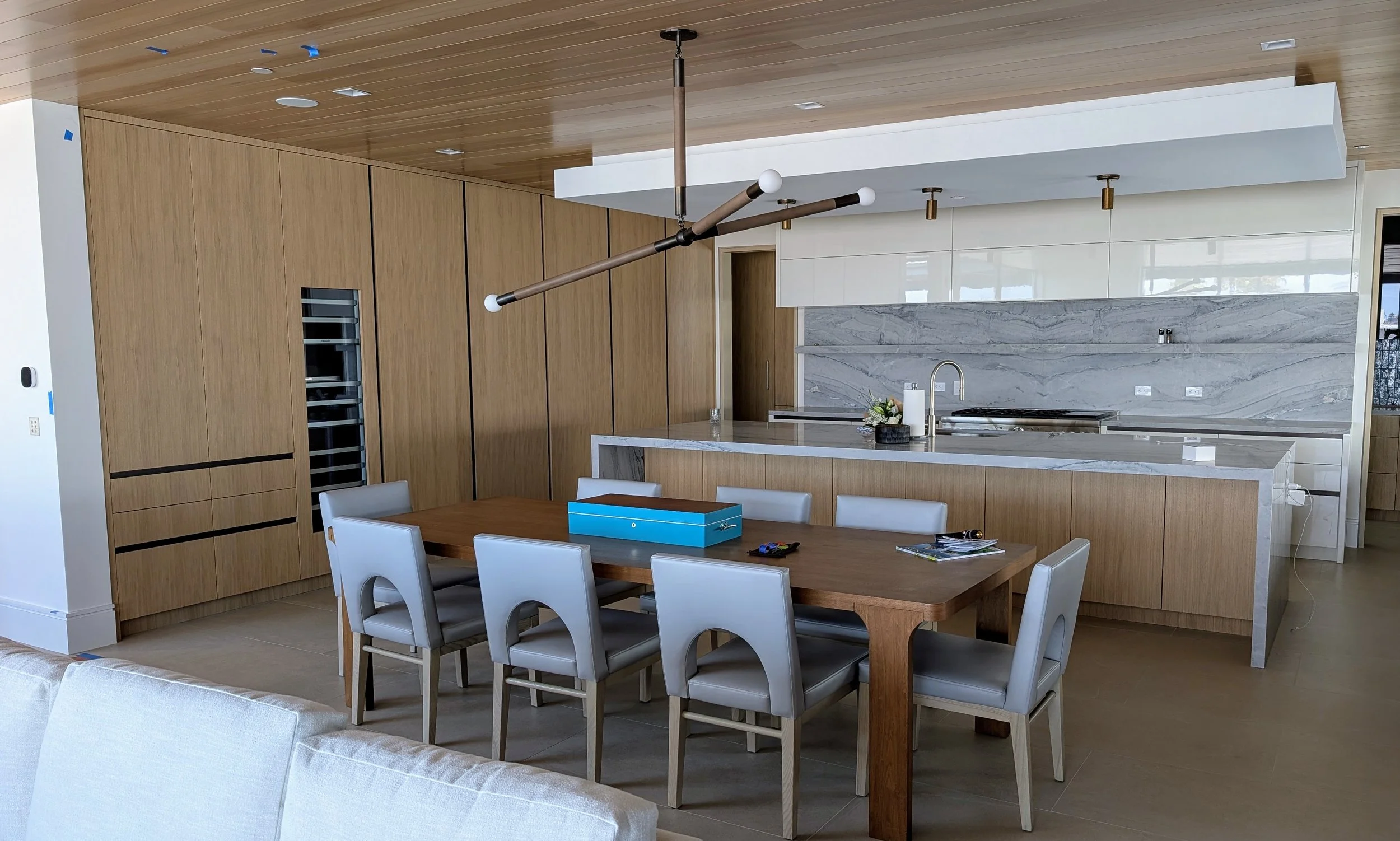 Modern open-plan kitchen with wooden cabinets, marble backsplash, and a kitchen island. Dining table with white chairs is in the foreground, and a contemporary chandelier hangs from the wooden ceiling.