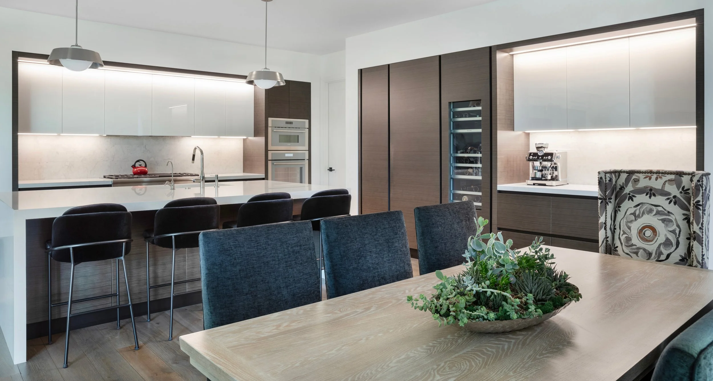 Modern kitchen with white and wooden cabinets, black bar stools, kitchen appliances, and a dining table with a plant centerpiece.