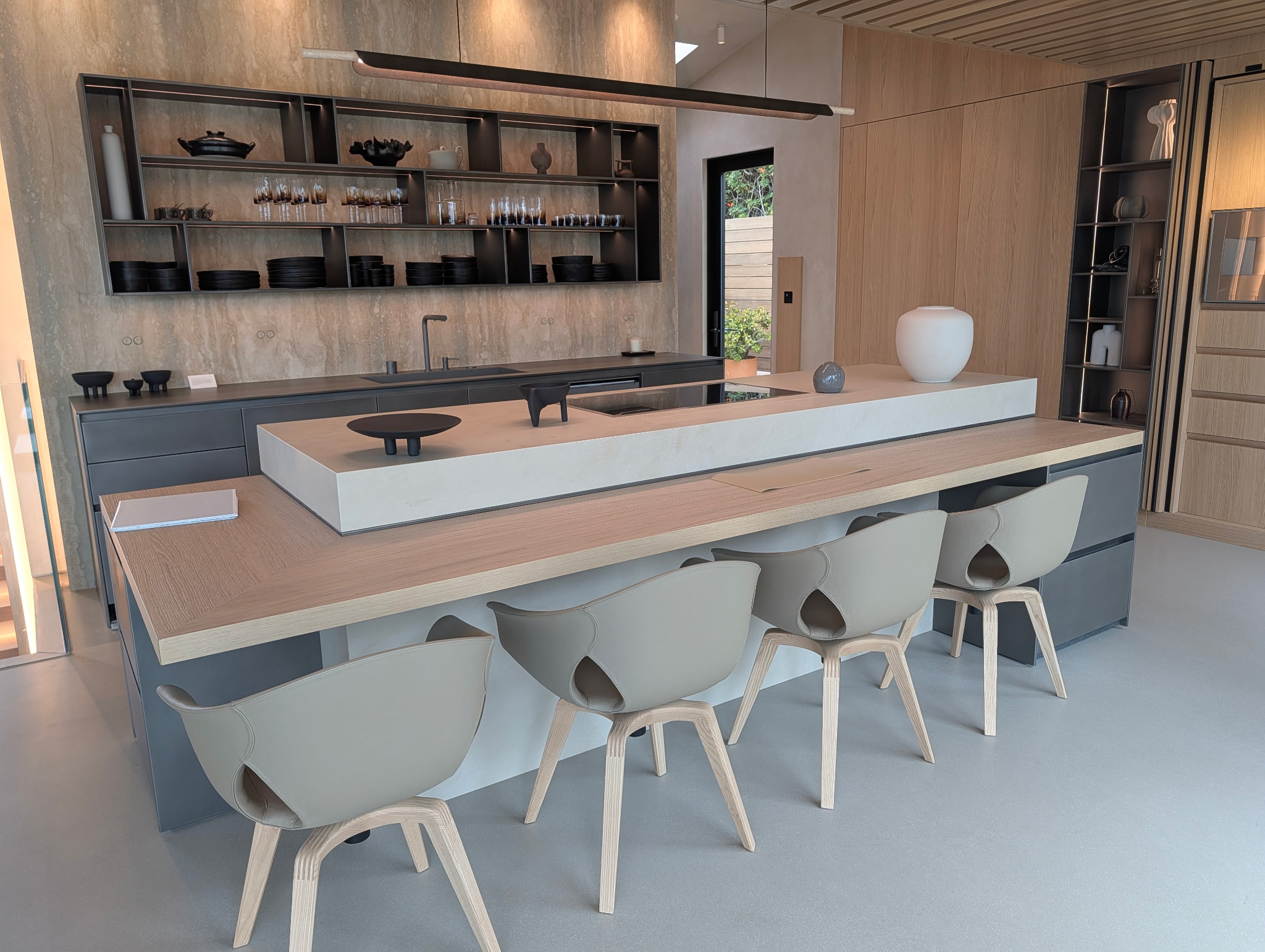 Modern kitchen with a large island, light-colored chairs, open shelving with black dishes and decorative items, large window with greenery outside.