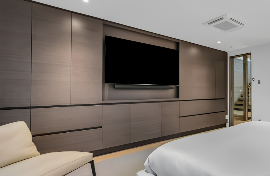 Modern hotel room with a flat-screen TV on a wooden wall, a beige chair, and a bed with white bedding. There is a doorway with stairs in the background.
