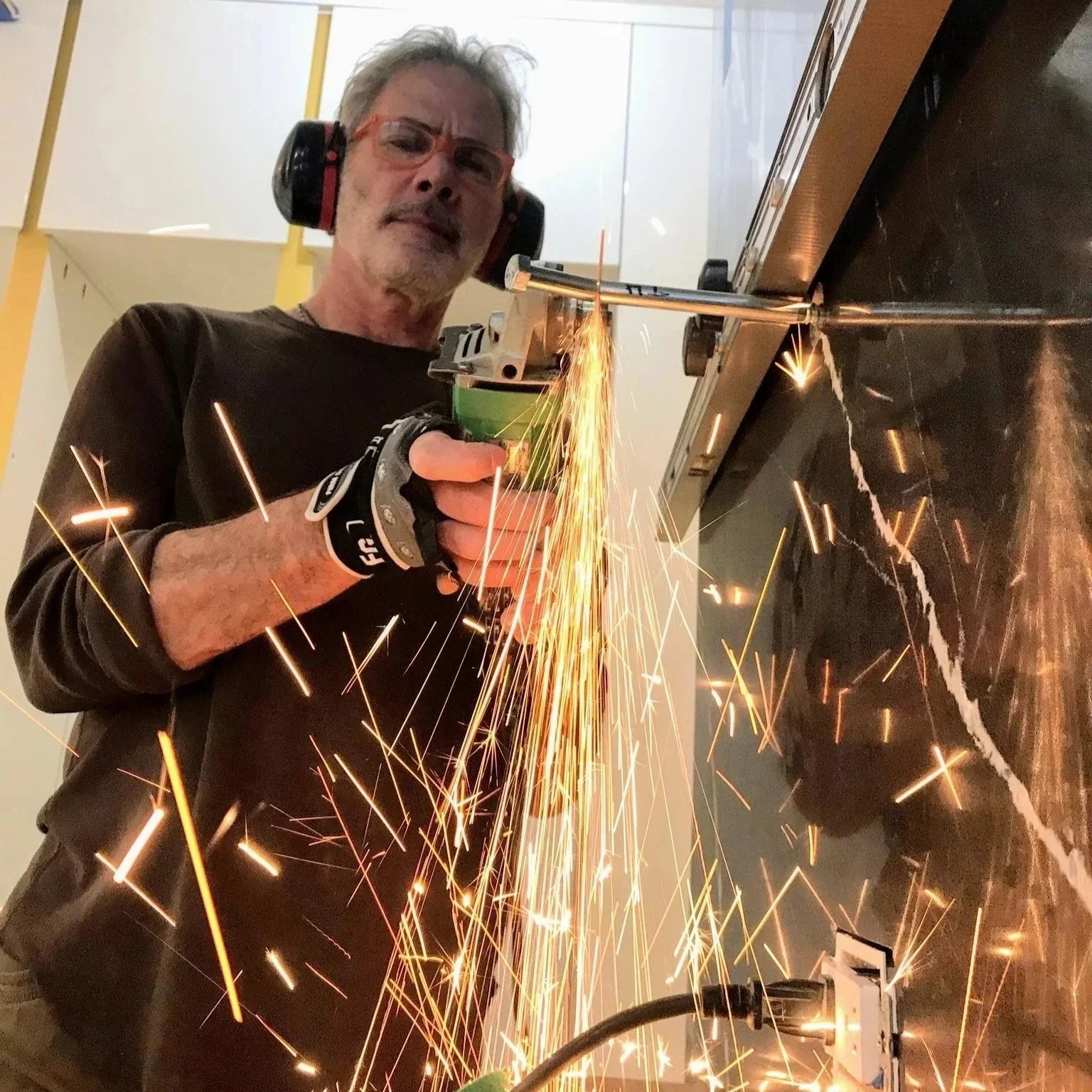A man with glasses and headphones using a handheld tool to cut or weld metal, creating bright sparks in a workshop or industrial setting.