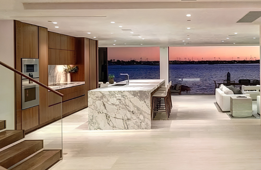 Interior view of a modern kitchen and living area with a large marble island, bar stools, built-in appliances, and a waterfront view with a sunset over the water.