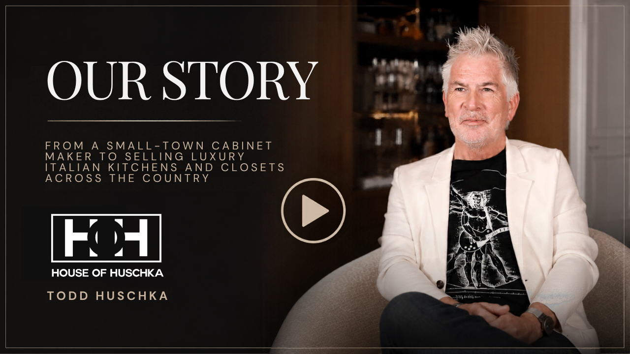 A man with gray hair and a beard, wearing a white blazer and a black graphic t-shirt, sitting on a beige couch with a dark background, promotional text for a show titled 'Our Story' with the logo for House of Huschka and Todd Huschka.