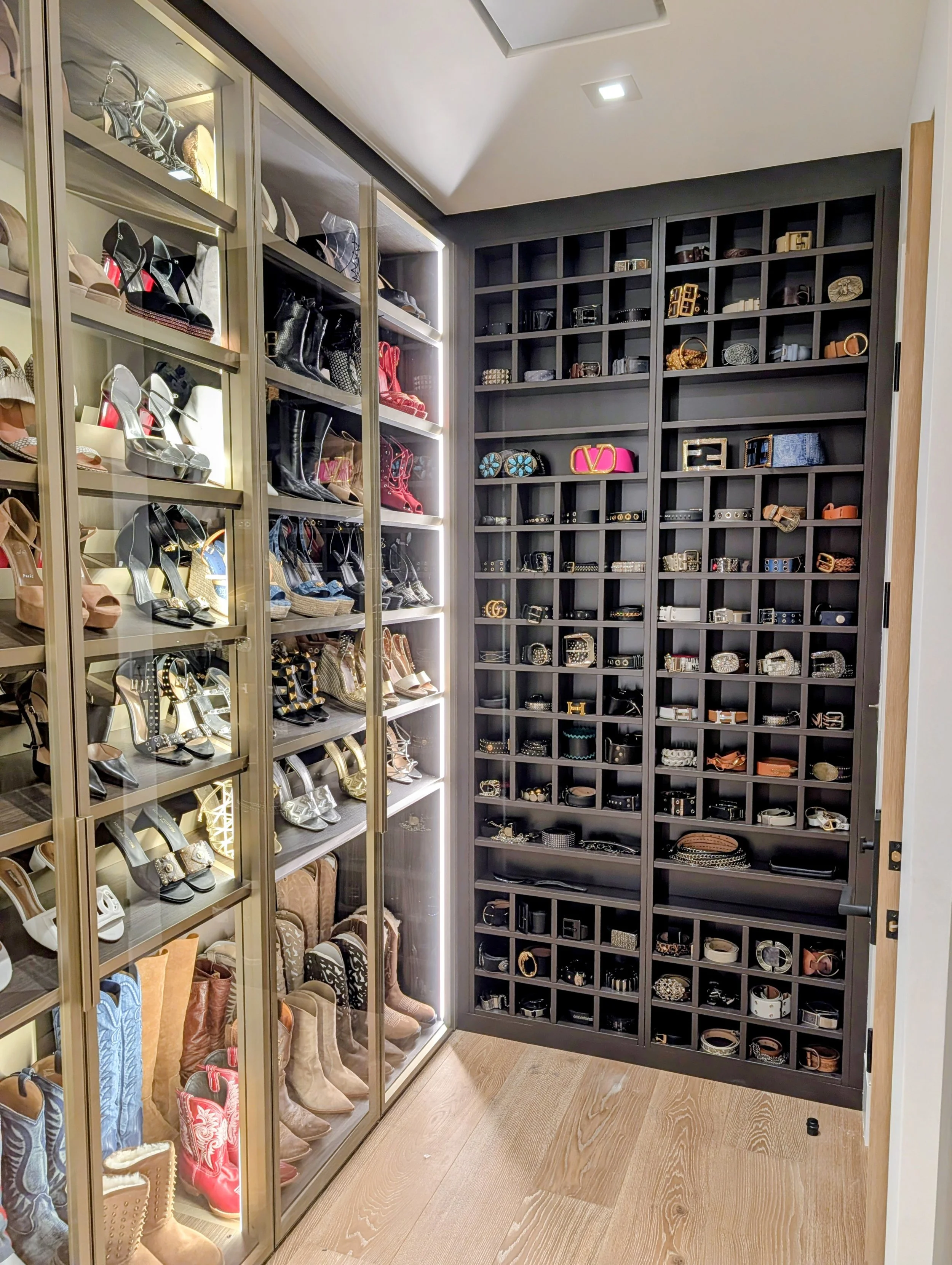 A walk-in closet with glass shelves on the left displaying various styles of women's shoes, including high heels and boots, and dark cubby shelves on the right storing belts, handbags, and accessories.