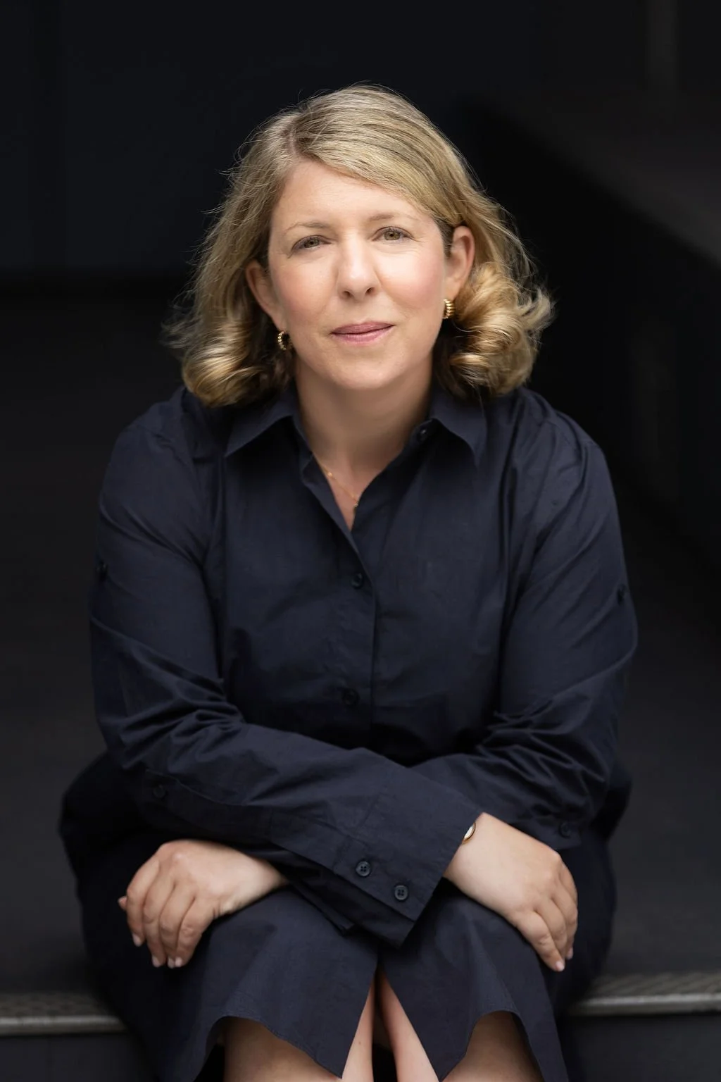A woman with blonde hair wearing a navy blue shirt, sitting against a dark background.