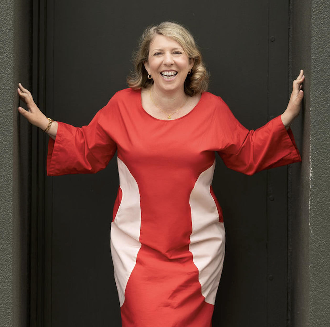 A smiling woman in a red and white dress standing in front of a black door with her arms extended, holding the door frame.