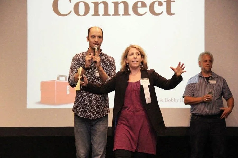 Three people standing in front of a screen with the word 'Connect' on it. The woman in the middle is speaking into a microphone and gesturing with her hand, while the two men on either side of her listen, one holding a drink and the other with a hand in his pocket.