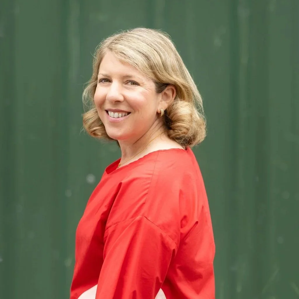 A smiling woman with shoulder-length blonde hair wearing a red top, standing in front of a green background.