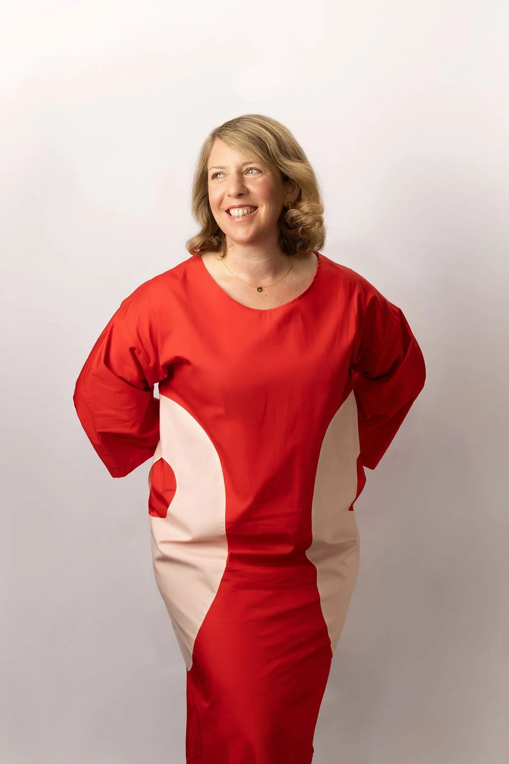 A woman with shoulder-length wavy blonde hair wearing a red and white dress, smiling and looking to her left, standing against a plain light gray background.