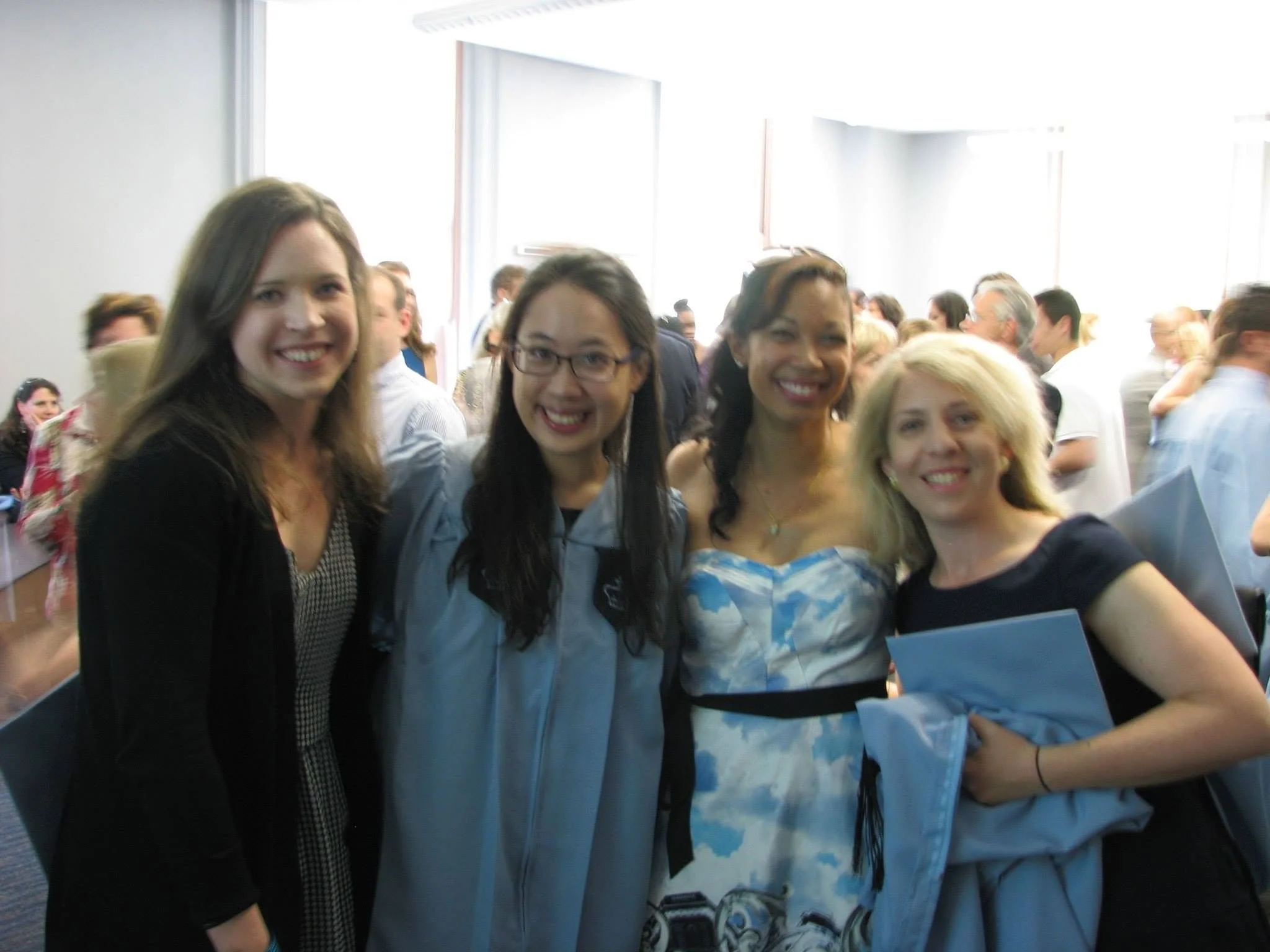 Four women smiling and posing for a photo in a crowded indoor setting, with many people in the background.