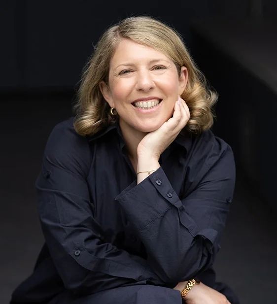 A woman with blonde hair smiling, wearing a dark blue shirt, resting her chin on her hand.