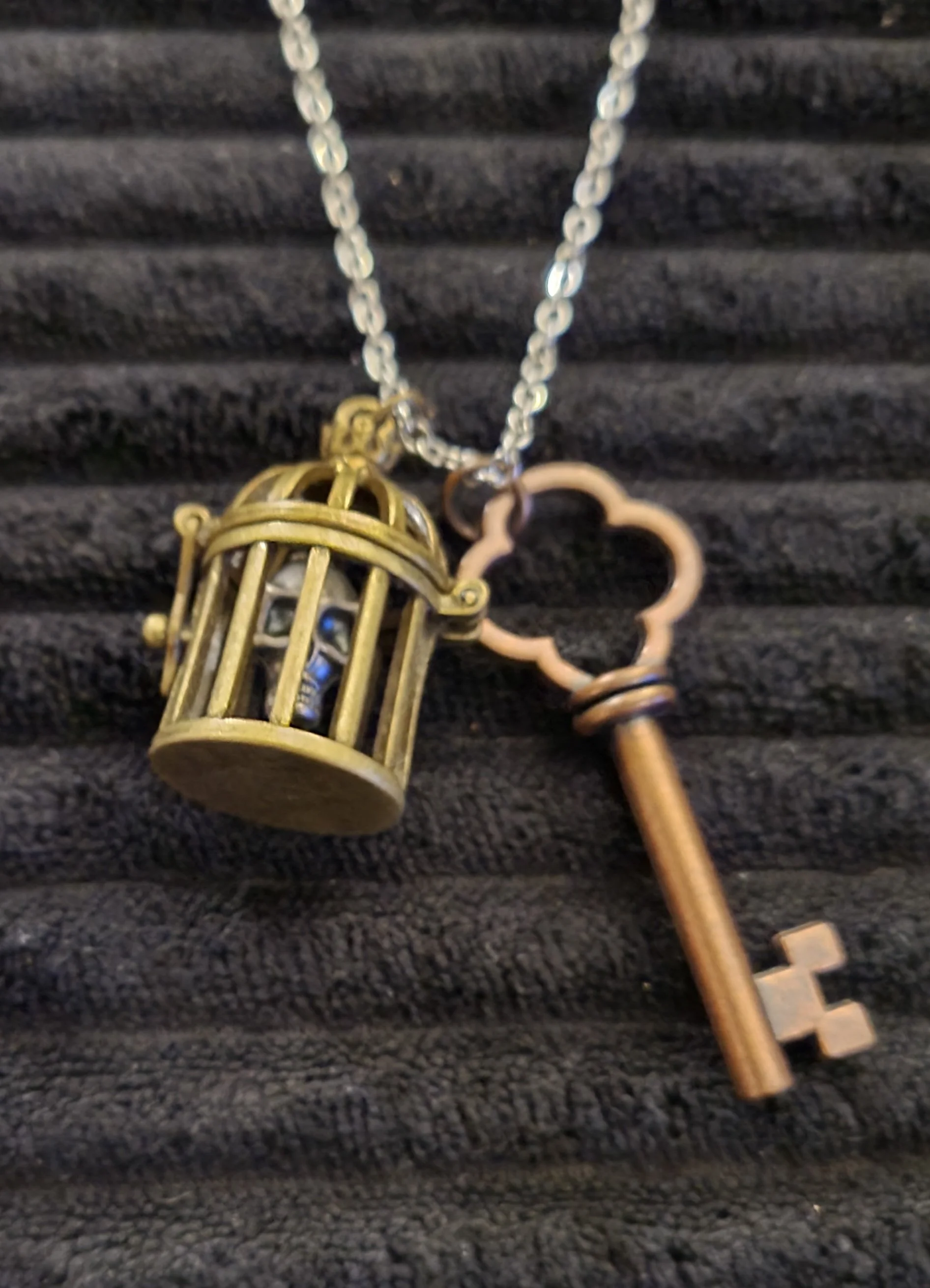 Up-close picture of a necklace with a key charm and a birdcage charm that has a skull charm inside it.