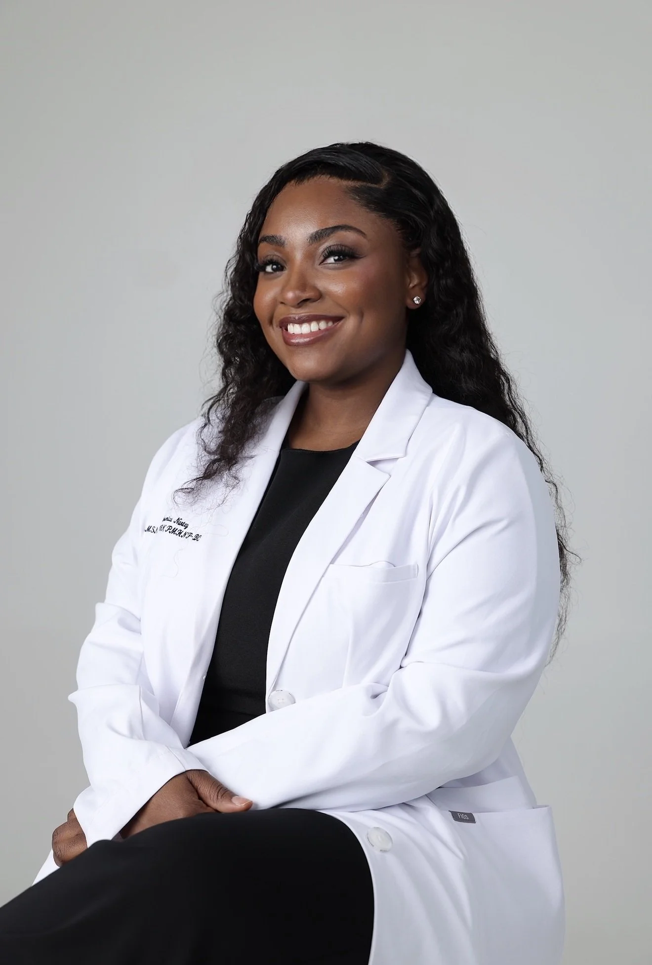 Portrait of a smiling African American woman with dark, curly hair, wearing a white medical coat, sitting against a plain gray background.
