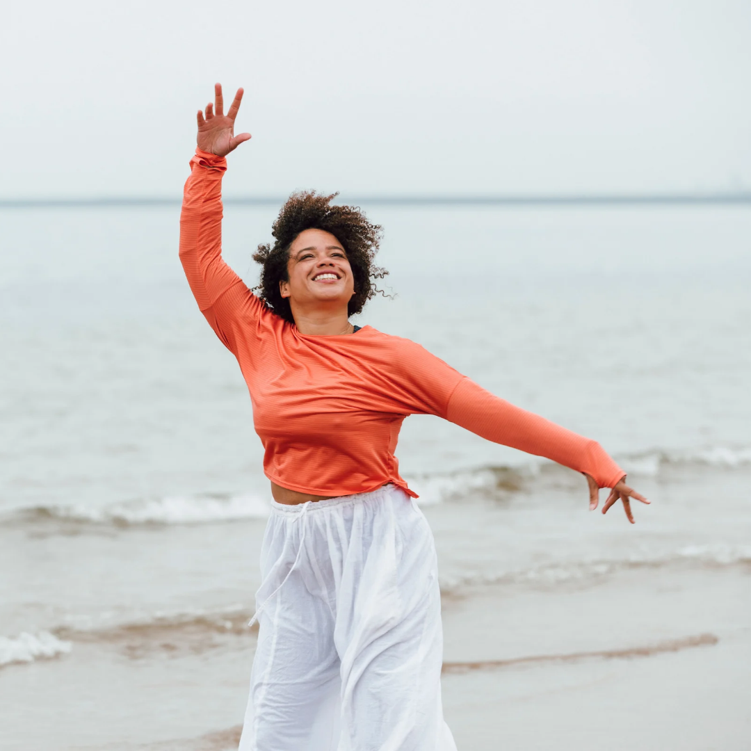 Woman smiling and dancing on the beach near the water, wearing an orange long sleeve top and white pants.
