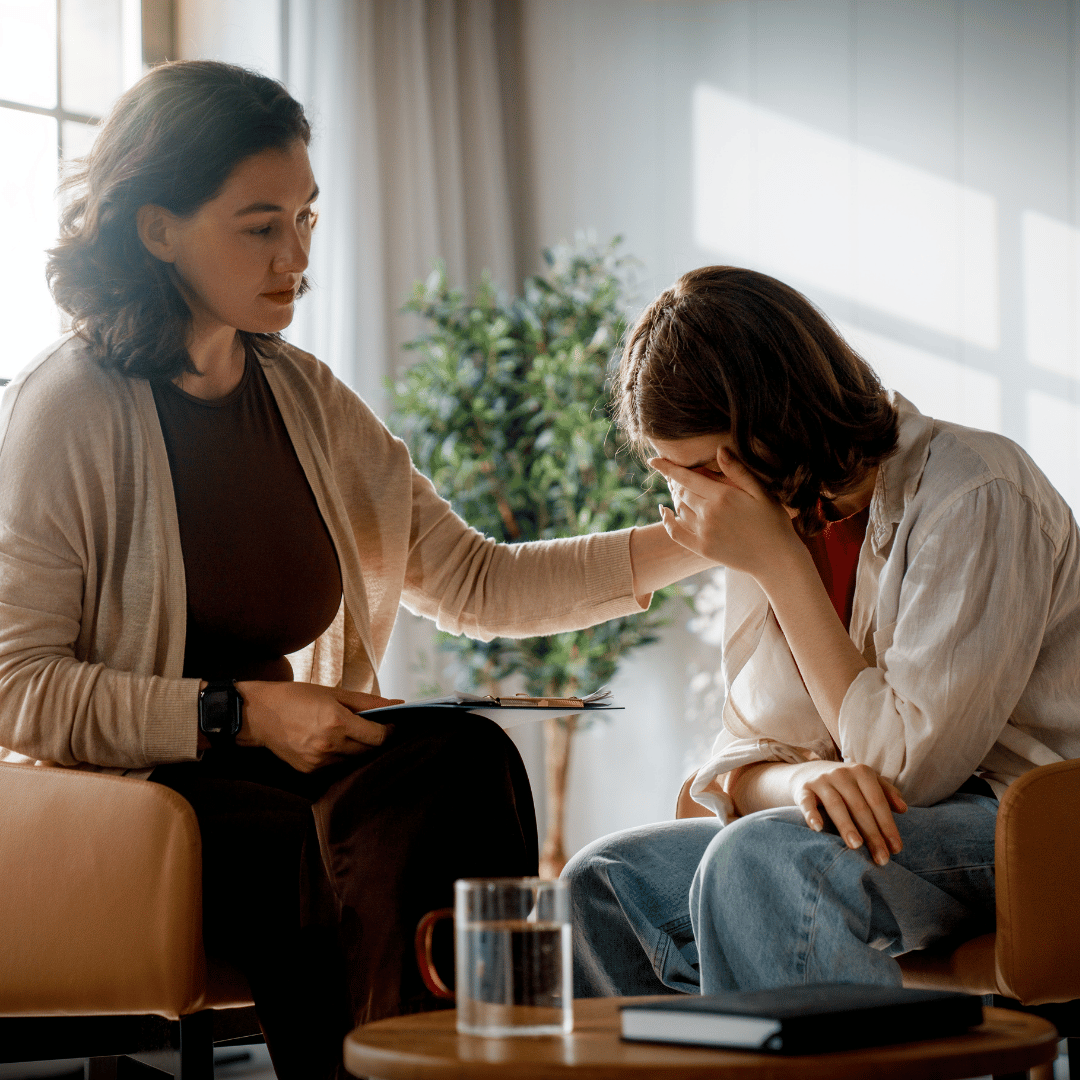 A woman comforting another woman who is sitting with her head in her hand, in what appears to be a therapy or counseling session in a bright room.