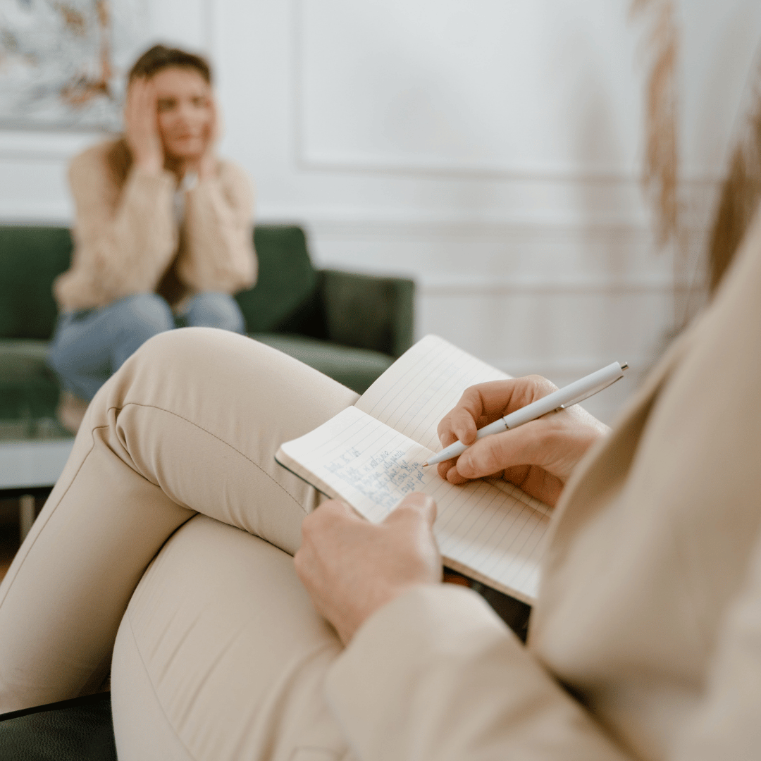 Therapist taking notes during a session with a woman sitting on a green couch, who appears upset.