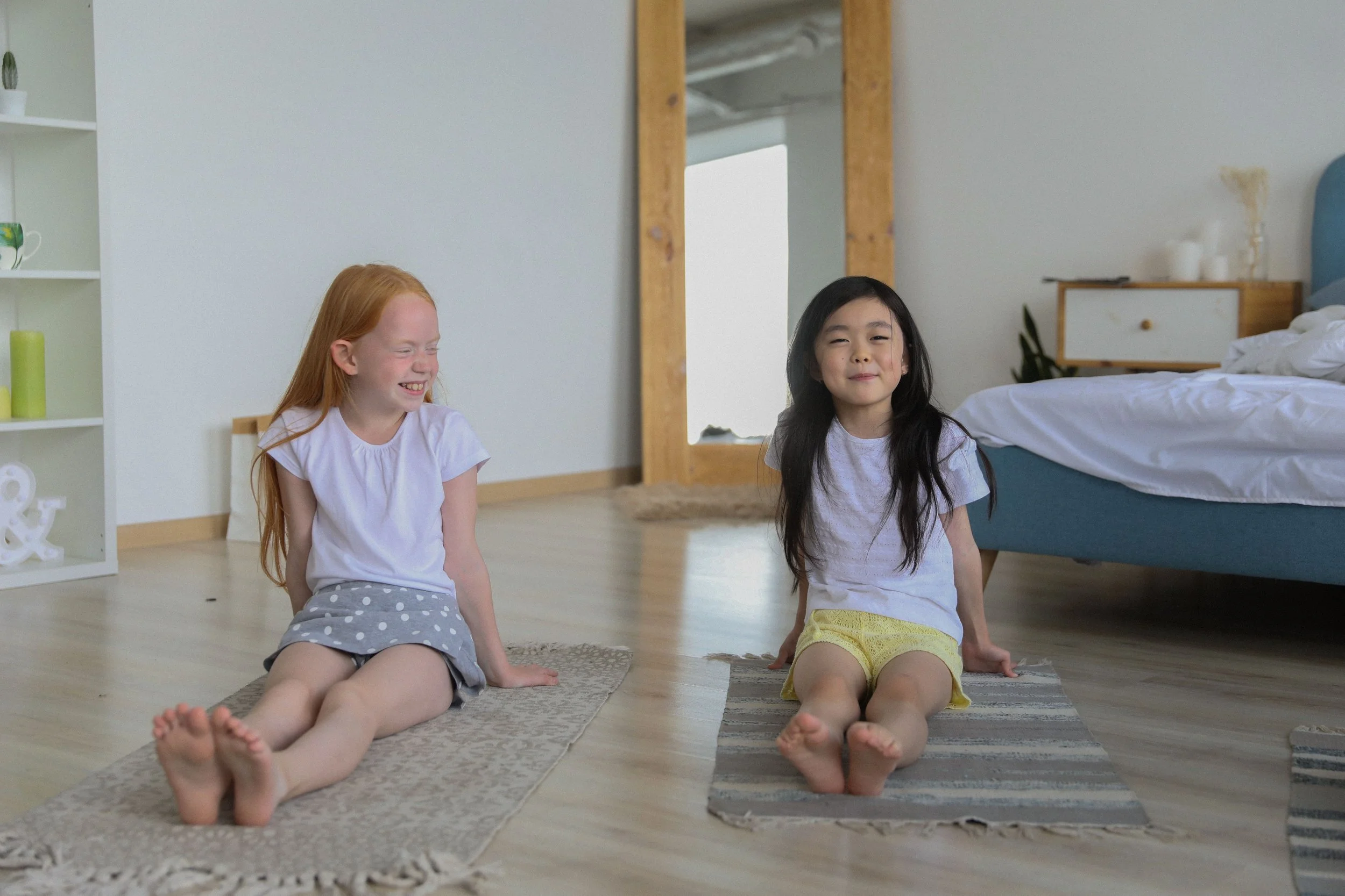 Two young girls sitting on the floor in a bedroom, smiling and relaxing, with bed and shelves in the background.