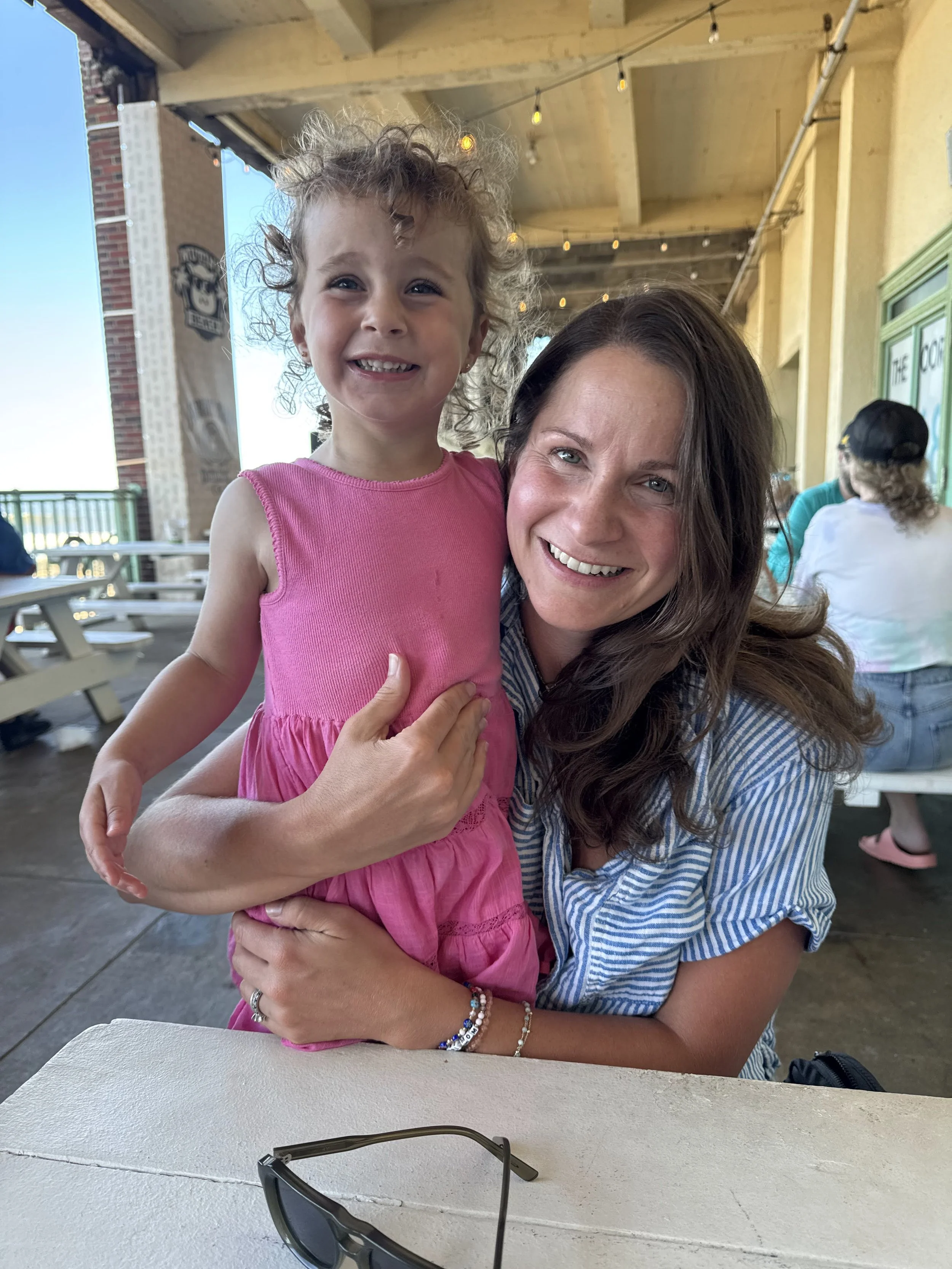 A woman with long brown hair smiling and holding a young girl with curly hair, wearing a pink dress, at an outdoor dining area with string lights and a wooden ceiling.