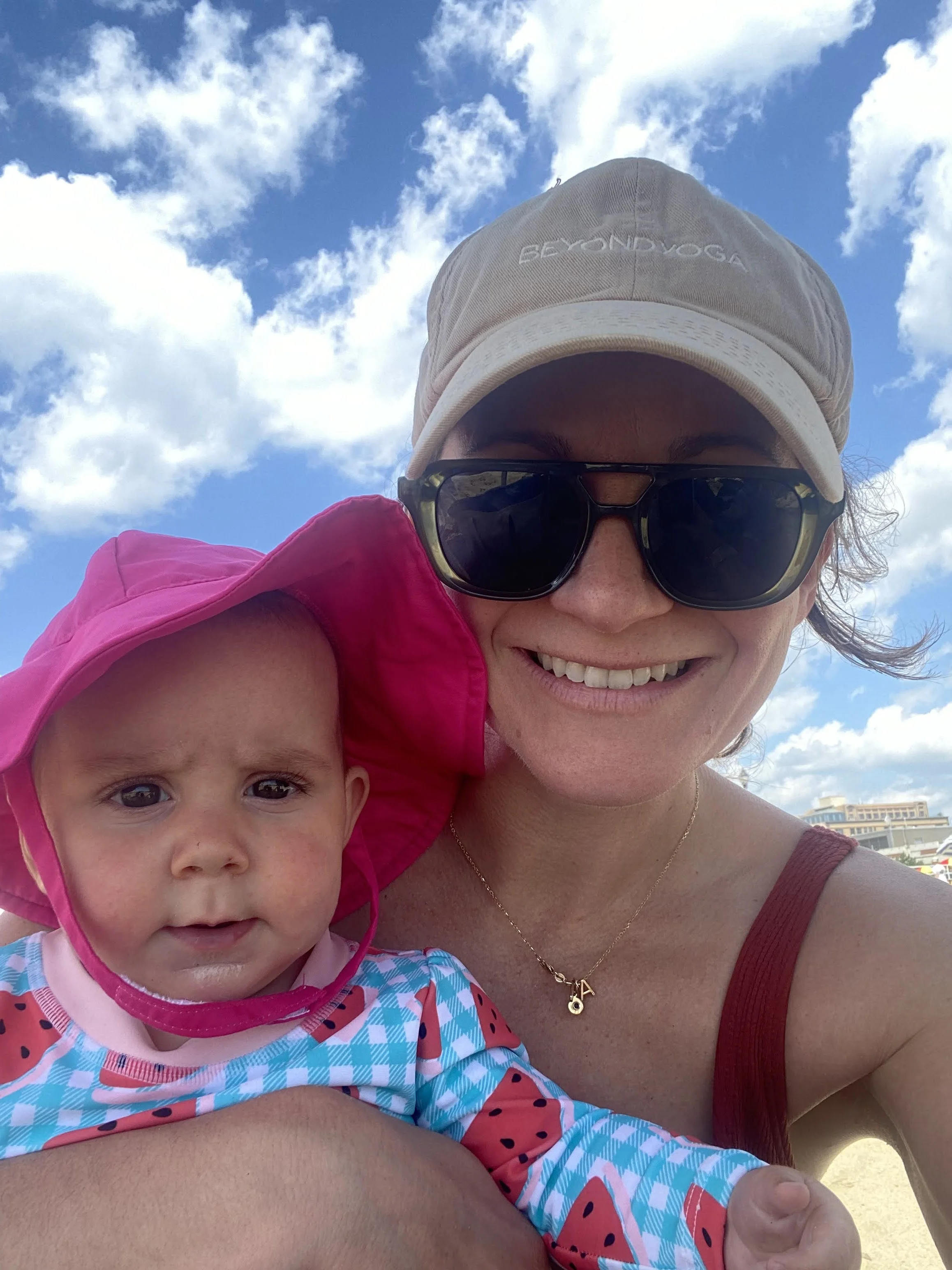 A woman wearing a white cap, sunglasses, and a red tank top taking a selfie with a happy baby girl wearing a pink, blue, and white patterned dress and a pink hooded jacket, outdoors under a blue sky with white clouds.