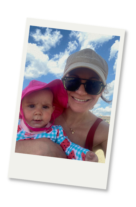 A woman with sunglasses and a beige cap holding a young child in a pink hat and colorful outfit, outdoors with a blue sky and clouds in the background.