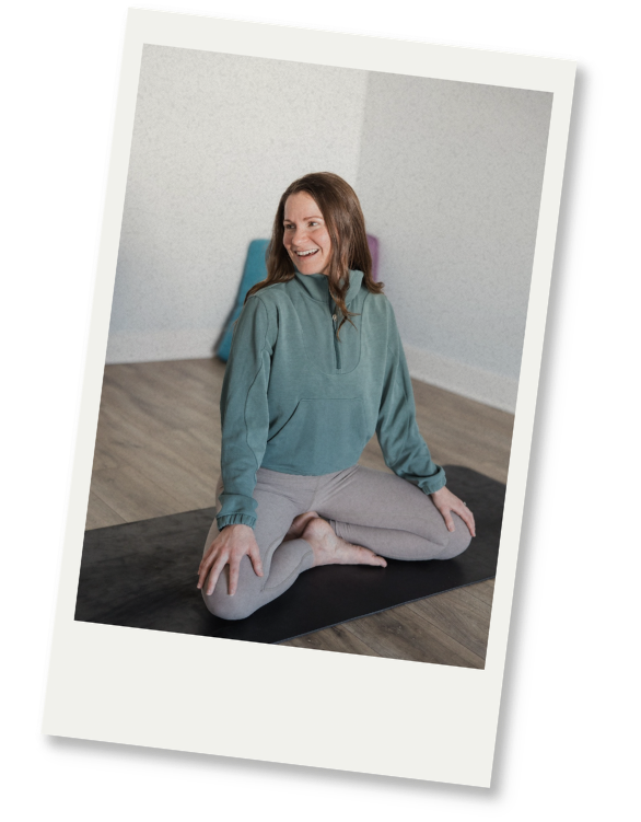 A woman with long brown hair sitting cross-legged on a yoga mat, smiling and looking to the side, dressed in a light blue hoodie and gray leggings, in a room with light-colored walls and wooden flooring.