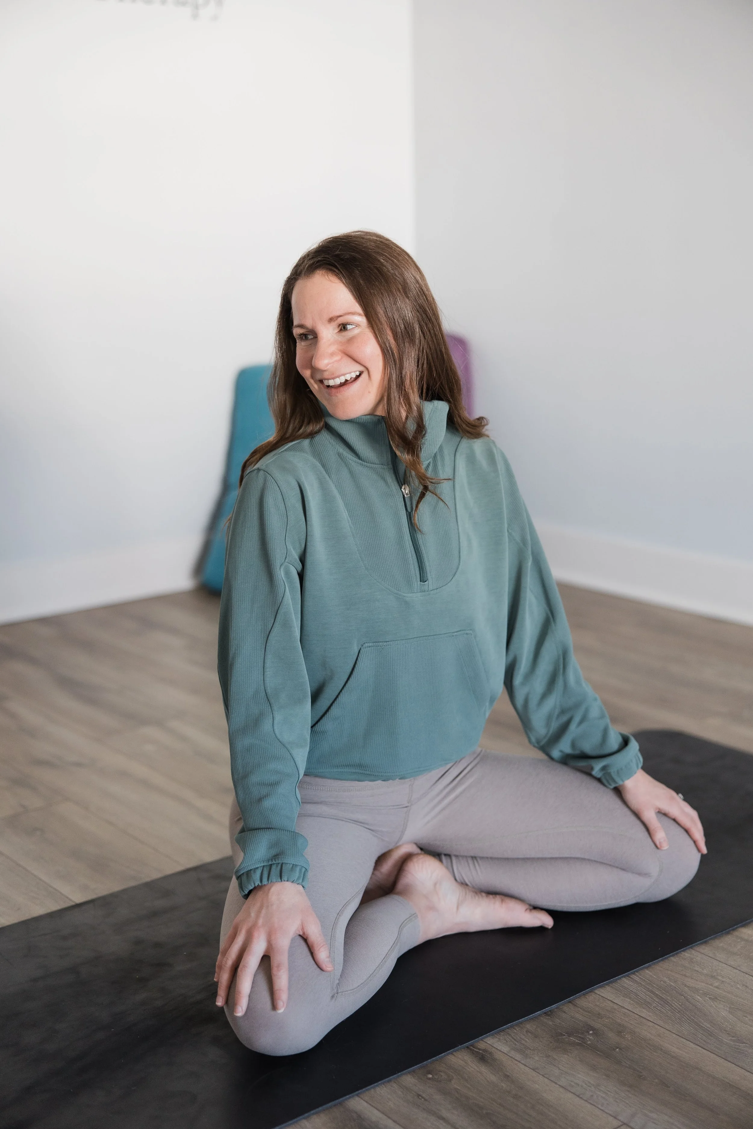 A woman practicing yoga indoors on a black yoga mat. She is wearing a teal pullover and light gray leggings, sitting in a cross-legged pose, with one hand resting on her knee and the other on the floor. She is smiling and looking to her left.